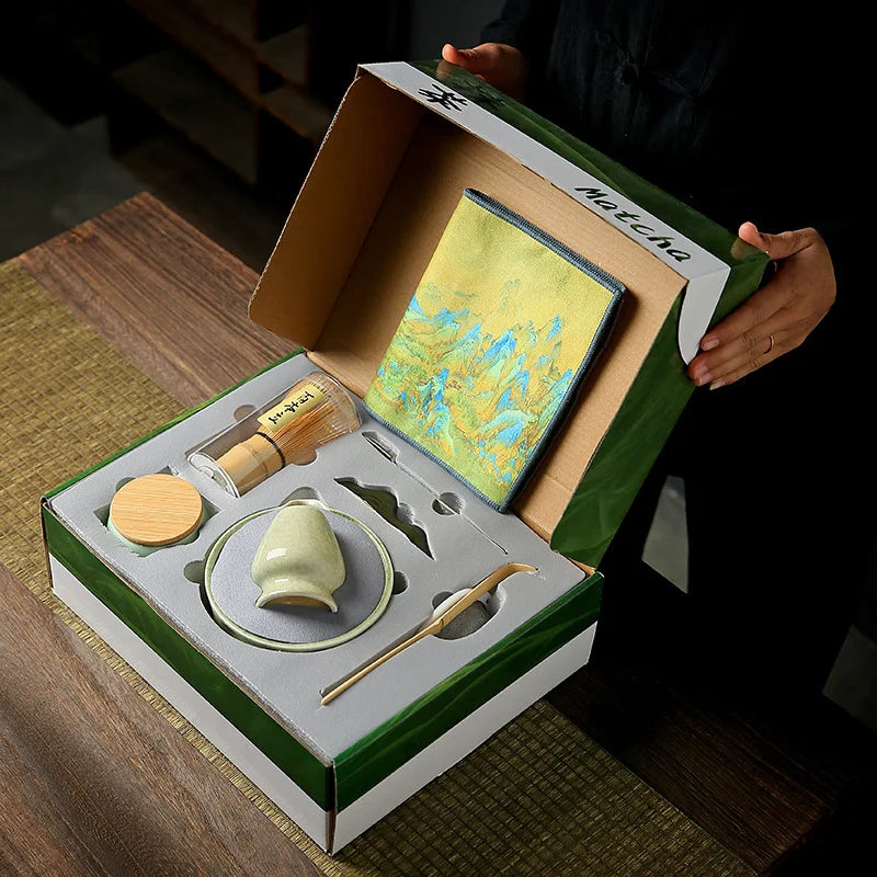 Coffret cadeau pour cérémonie du matcha, ouvert sur une table en bois, contenant un chasen (fouet en bambou), un chashaku (cuillère en bambou), un chawan (bol en céramique), un support pour chasen, une boîte à thé, un tamis et un tissu décoratif, élégamment rangés dans une mousse de protection.