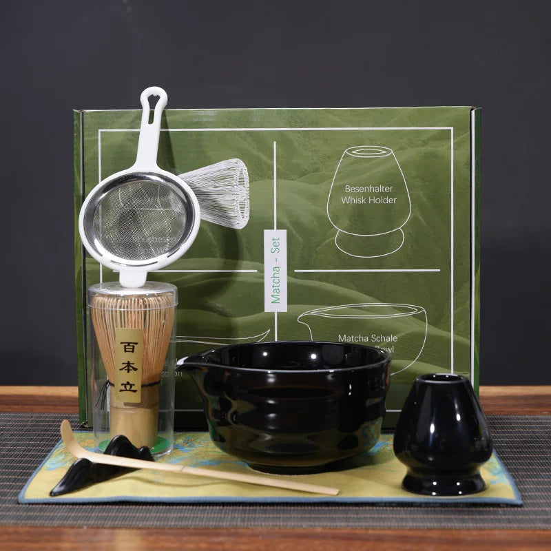 Ensemble de cérémonie du thé matcha aux tons noirs et naturels, exposé devant une boîte verte illustrée. Le set comprend un tamis en inox avec poignée blanche, un chasen (fouet en bambou clair) posé sur un support vert céladon, un chawan (bol à matcha) en céramique noir, une cuillère doseuse métallique, un pot à thé noir pâle avec couvercle en bois clair, et un tissu décoratif vert anis aux motifs bleus et dorés.