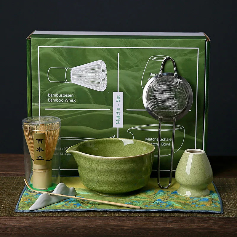 Ensemble de cérémonie du thé matcha aux tons verts et naturels, exposé devant une boîte verte illustrée. Le set comprend un tamis en inox avec poignée blanche, un chasen (fouet en bambou clair) posé sur un support vert, un chawan (bol à matcha) en céramique vert, une cuillère doseuse métallique, un pot à thé vert avec couvercle en bois clair, et un tissu décoratif vert anis aux motifs bleus et dorés.