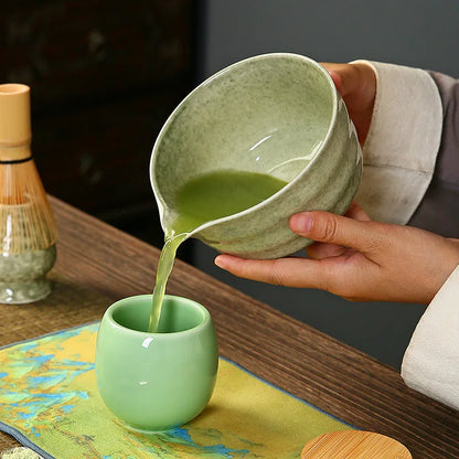 Un chawan traditionnel en céramique verse doucement du matcha mousseux dans un petit bol vert, laissant couler un filet vert vif et onctueux.