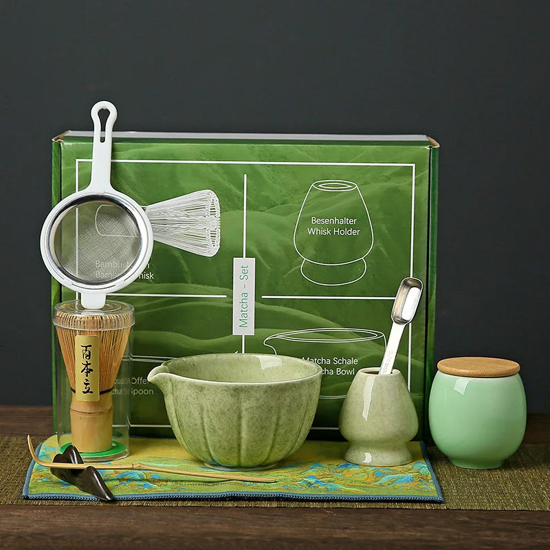 Ensemble de cérémonie du thé matcha aux tons verts et naturels, exposé devant une boîte verte illustrée. Le set comprend un tamis en inox avec poignée blanche, un chasen (fouet en bambou clair) posé sur un support vert céladon, un chawan (bol à matcha) en céramique vert jade, une cuillère doseuse métallique, un pot à thé vert pâle avec couvercle en bois clair, et un tissu décoratif vert anis aux motifs bleus et dorés.