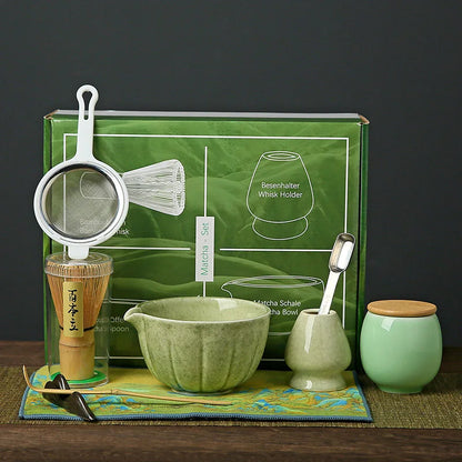Ensemble de cérémonie du thé matcha aux tons verts et naturels, exposé devant une boîte verte illustrée. Le set comprend un tamis en inox avec poignée blanche, un chasen (fouet en bambou clair) posé sur un support vert céladon, un chawan (bol à matcha) en céramique vert jade, une cuillère doseuse métallique, un pot à thé vert pâle avec couvercle en bois clair, et un tissu décoratif vert anis aux motifs bleus et dorés.